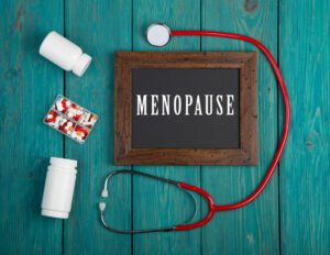 A blackboard displaying the word Menopause, accompanied by a stethoscope and pills, set against a blue wooden background.