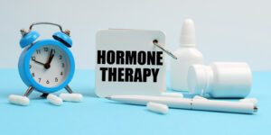 On a blue surface, there are pills, an alarm clock, and a notebook labeled HORMONE THERAPY.
