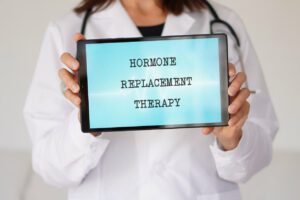 The phrase HORMONE REPLACEMENT THERAPY is displayed on a tablet held by a female physician.