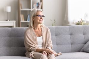 A thoughtful retired woman, considering hormone replacement therapy (HRT), sitting on a sofa and gazing off to the side with a gentle smile.